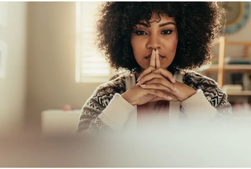 Women in a cozy sweater with fingers interlaced, both index fingers across her lips looking directly at the camera 