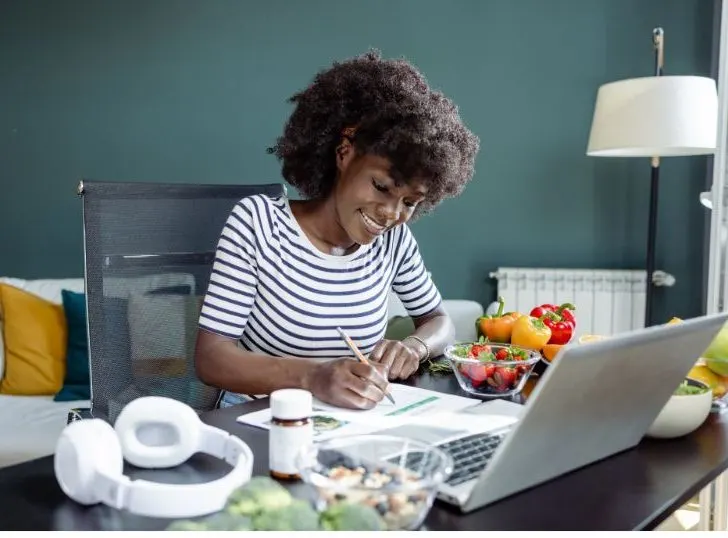 A beautiful, smiling African wearing a black and white shirt sits in front of the laptop, planning and calculating her daily meal.
