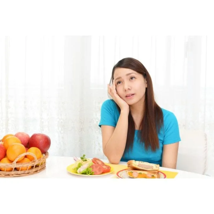 A woman in a blue t-shirt sits in front of food, pondering what to eat today.