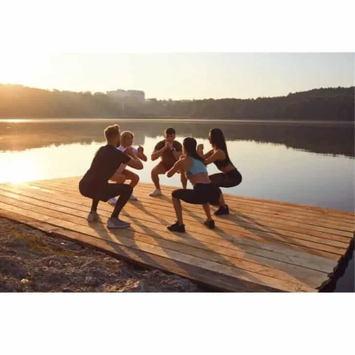 A group of men and women are working out, smiling, and doing squats at sunset.
